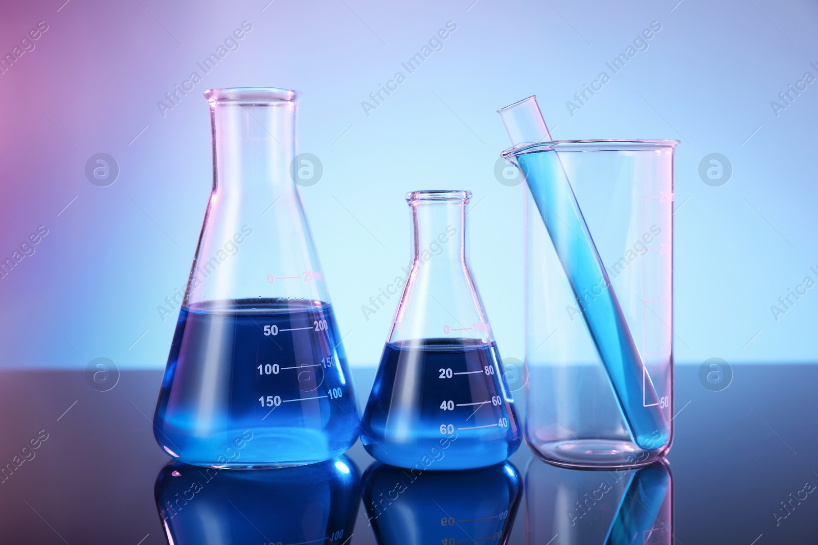 Photo of Different glassware with blue liquid on mirror surface against color background