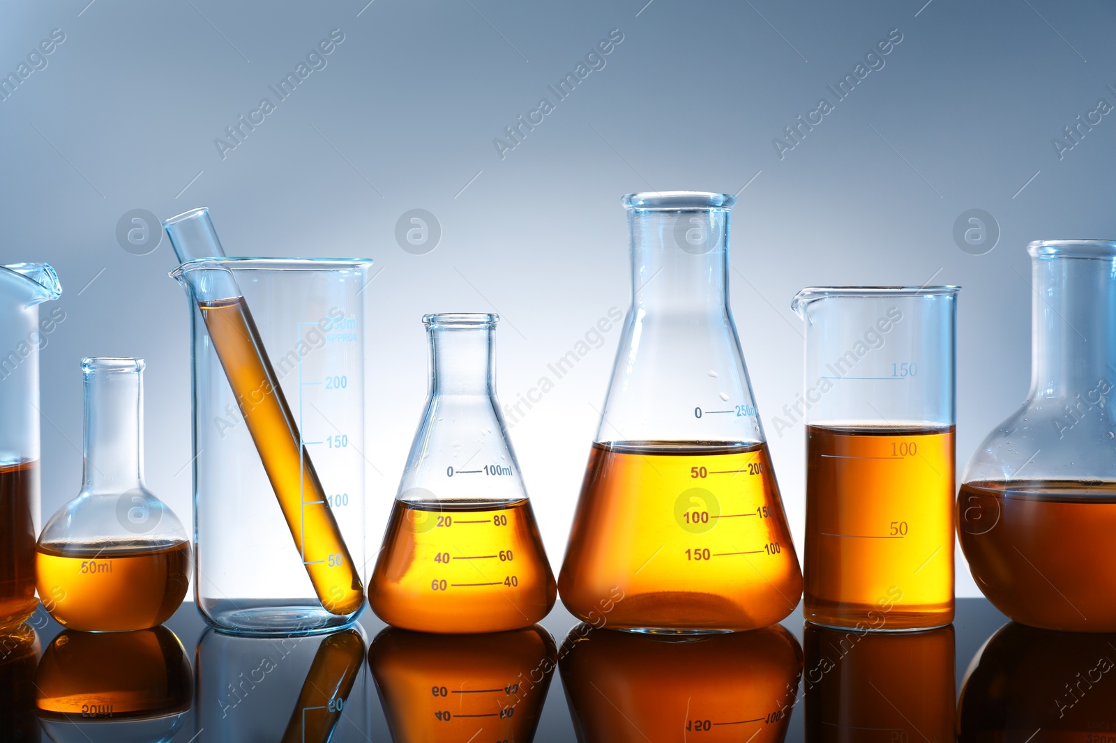 Photo of Different glassware with orange liquid on mirror surface against grey background