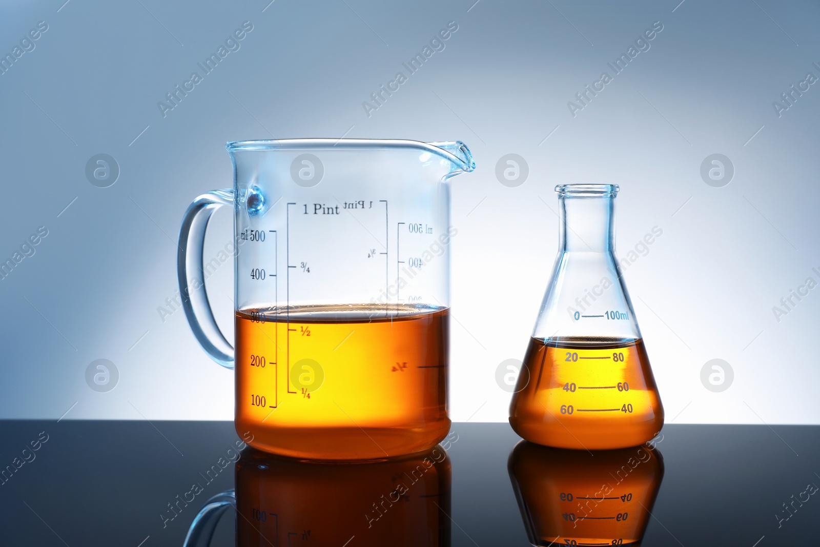 Photo of Glass flask and beaker with orange liquid on mirror surface against grey background