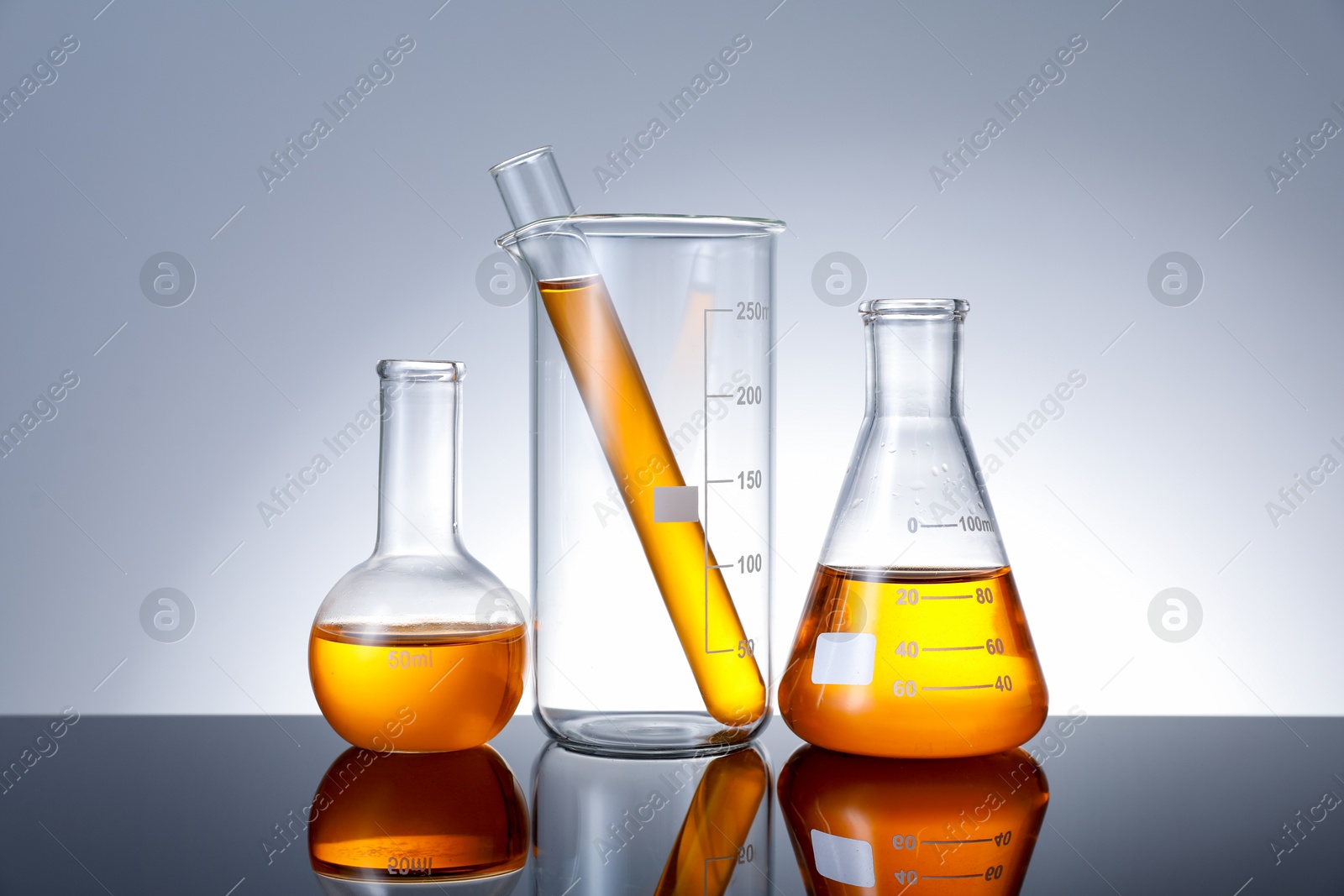Photo of Different glassware with orange liquid on mirror surface against grey background