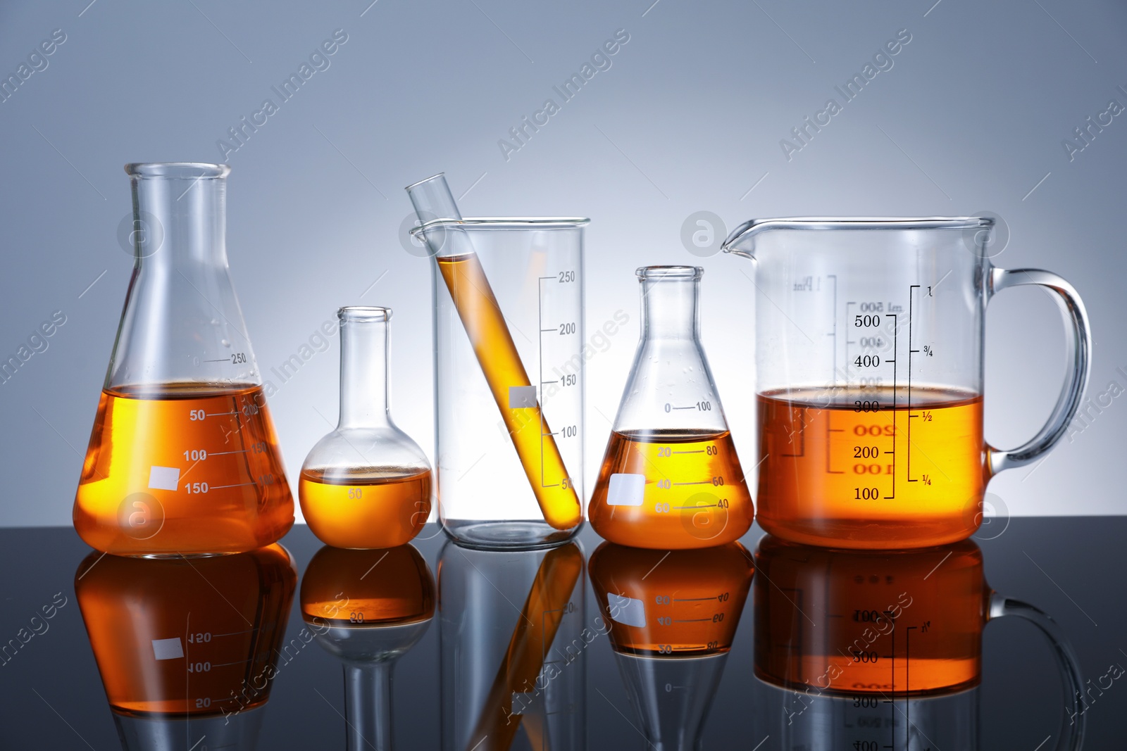 Photo of Different glassware with orange liquid on mirror surface against grey background