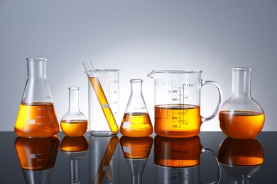 Photo of Different glassware with orange liquid on mirror surface against grey background