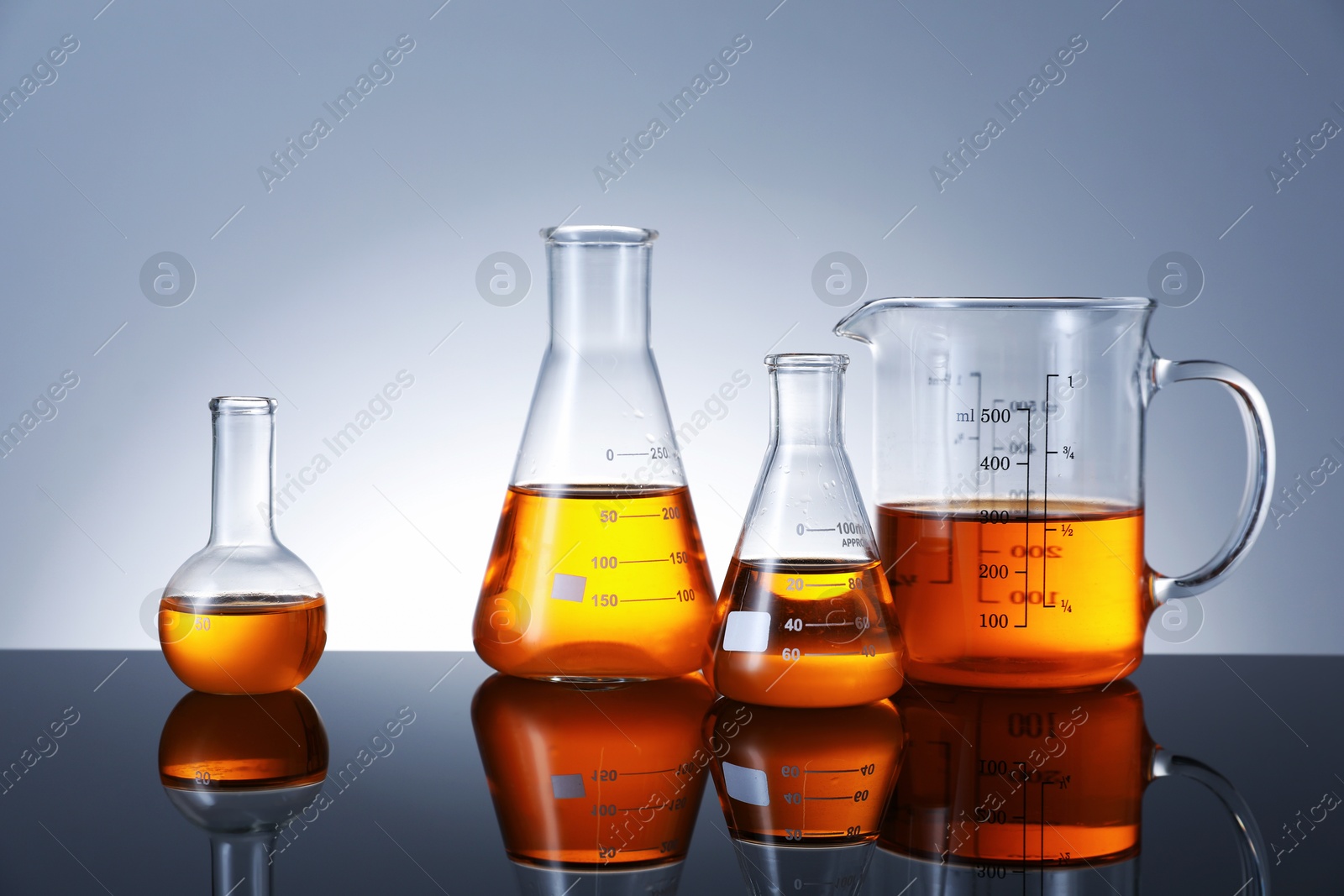 Photo of Different glassware with orange liquid on mirror surface against grey background