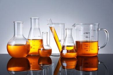 Photo of Different glassware with orange liquid on mirror surface against grey background