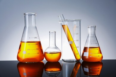 Photo of Different glassware with orange liquid on mirror surface against grey background