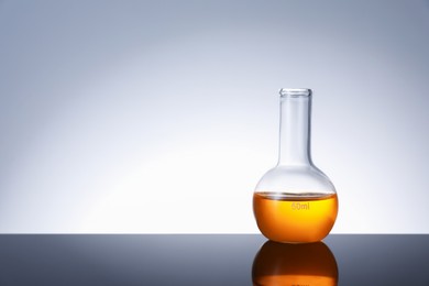 Photo of Glass flask with orange liquid on mirror surface against grey background, space for text
