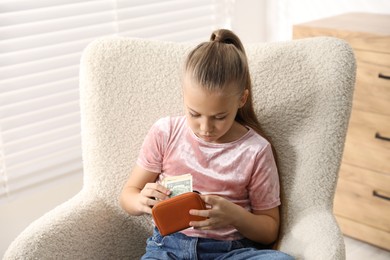 Photo of Little girl putting her pocket money into purse in armchair at home