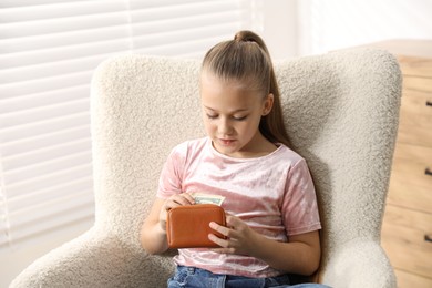 Photo of Little girl putting her pocket money into purse in armchair at home