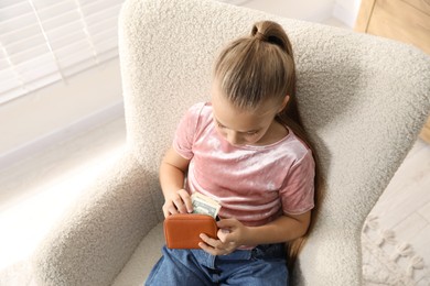 Photo of Little girl putting her pocket money into purse in armchair at home, above view