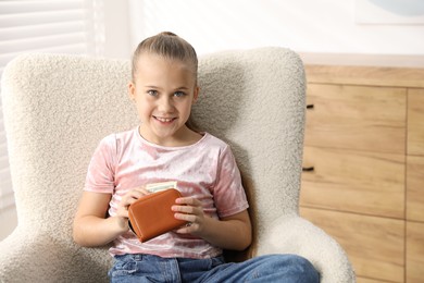 Photo of Little girl putting her pocket money into purse in armchair at home