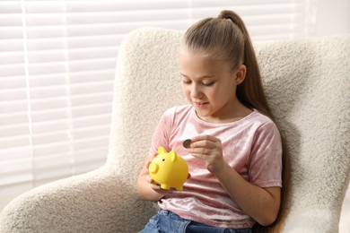 Photo of Little girl putting her pocket money into piggy bank in armchair at home