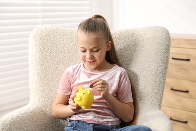 Photo of Little girl putting her pocket money into piggy bank in armchair at home