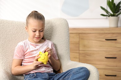 Photo of Little girl putting her pocket money into piggy bank in armchair at home