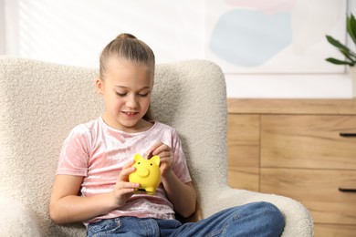 Photo of Little girl putting her pocket money into piggy bank in armchair at home