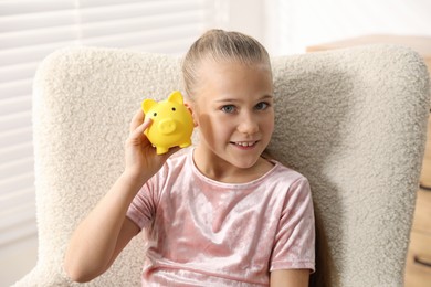 Photo of Pocket money. Little girl with piggy bank in armchair at home