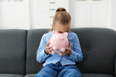 Photo of Pocket money. Little girl with piggy bank on sofa at home