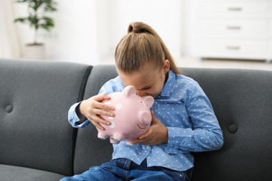 Photo of Pocket money. Little girl with piggy bank on sofa at home