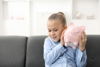 Photo of Pocket money. Little girl with piggy bank on sofa at home, space for text