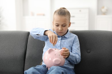 Photo of Little girl putting her pocket money into piggy bank on sofa at home