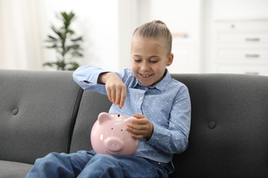 Photo of Little girl putting her pocket money into piggy bank on sofa at home