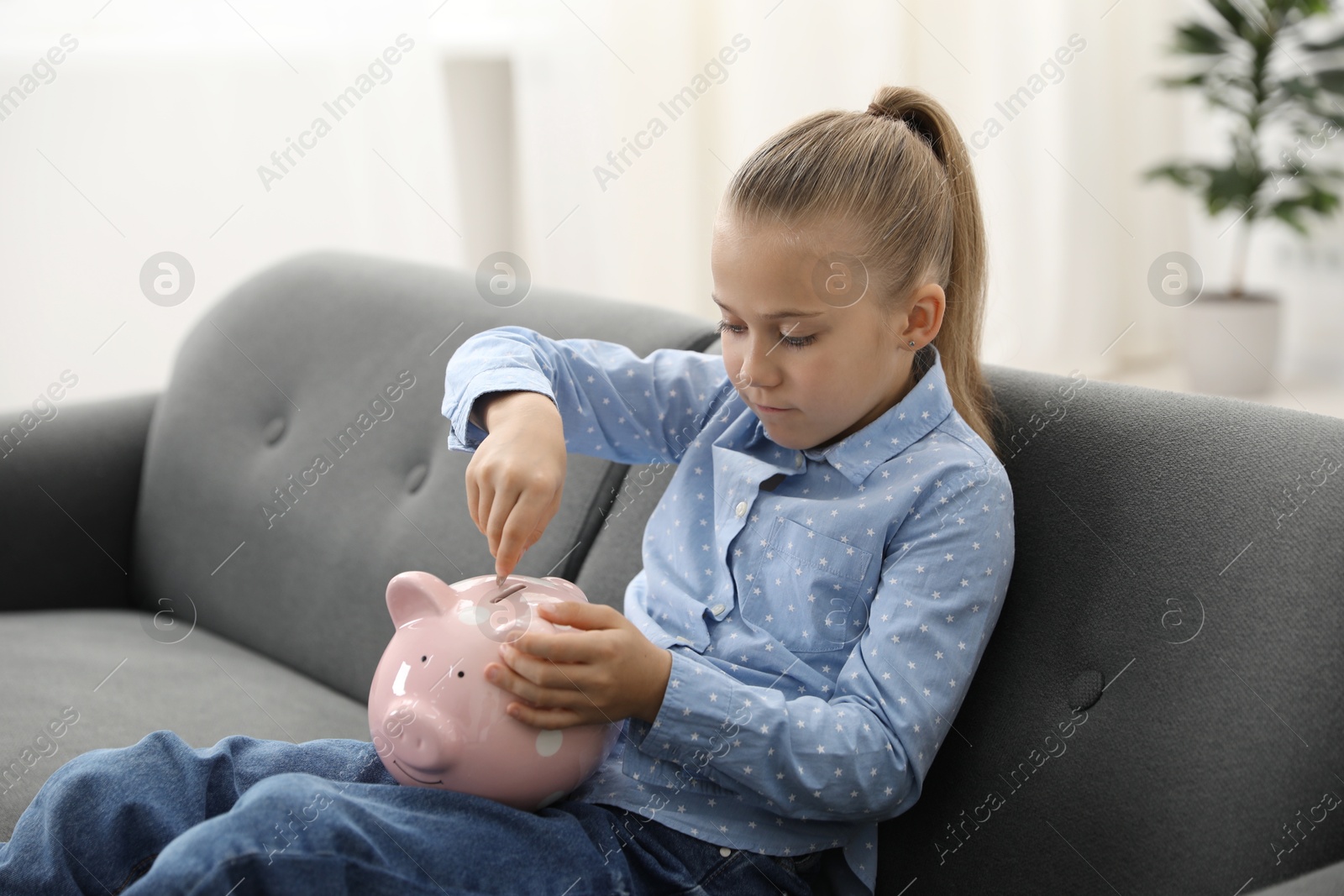Photo of Little girl putting her pocket money into piggy bank on sofa at home