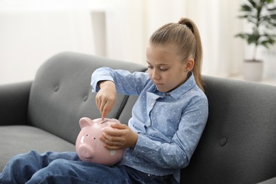 Photo of Little girl putting her pocket money into piggy bank on sofa at home
