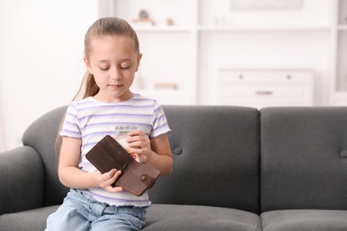 Photo of Little girl putting pocket money into purse on sofa at home, space for text