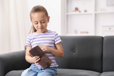 Photo of Little girl putting pocket money into purse on sofa at home, space for text