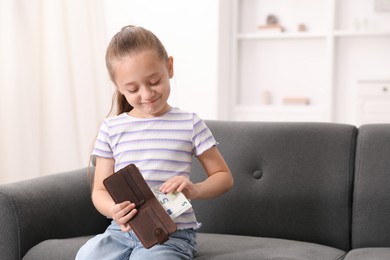Photo of Little girl putting pocket money into purse on sofa at home, space for text