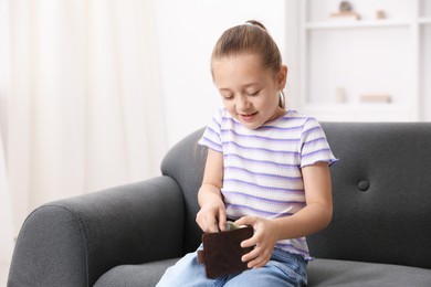 Photo of Little girl putting pocket money into purse on sofa at home