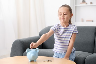 Photo of Little girl putting her pocket money into piggy bank at wooden table indoors