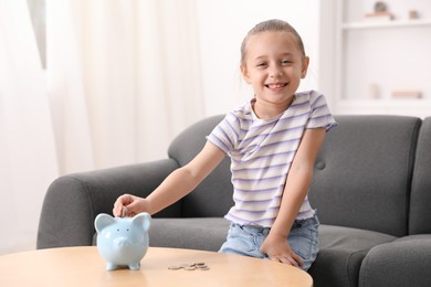 Photo of Little girl putting her pocket money into piggy bank at wooden table indoors