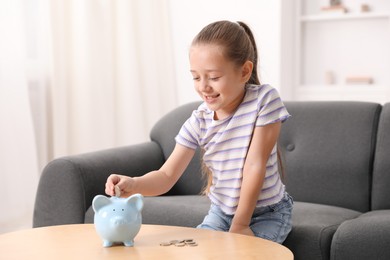 Photo of Little girl putting her pocket money into piggy bank at wooden table indoors