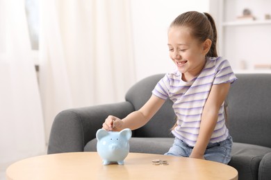 Photo of Little girl putting her pocket money into piggy bank at wooden table indoors