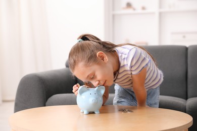 Photo of Little girl putting her pocket money into piggy bank at wooden table indoors