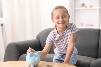 Photo of Little girl putting her pocket money into piggy bank at wooden table indoors