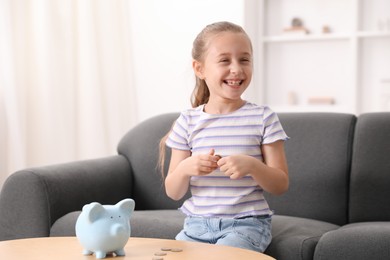 Photo of Pocket money. Little girl at wooden table with piggy bank and coins indoors