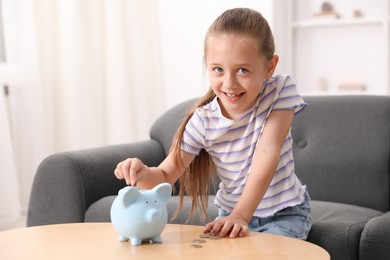 Photo of Little girl putting her pocket money into piggy bank at wooden table indoors