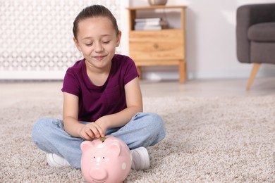Photo of Little girl putting her pocket money into piggy bank on floor at home, space for text