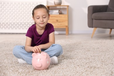 Photo of Little girl putting her pocket money into piggy bank on floor at home, space for text