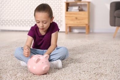 Photo of Little girl putting her pocket money into piggy bank on floor at home, space for text