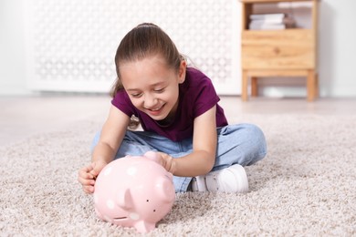 Photo of Little girl putting her pocket money into piggy bank on floor at home