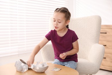 Photo of Little girl putting pocket money from broken piggy bank at wooden table indoors