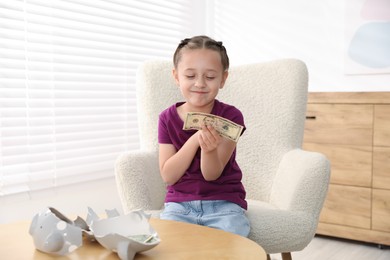 Photo of Little girl with pocket money in armchair at home