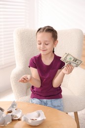 Photo of Little girl with pocket money in armchair at home