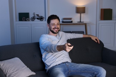 Photo of Happy man changing TV channels with remote control on sofa at home
