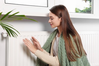 Photo of Woman in blanket warming hands on white heating radiator indoors