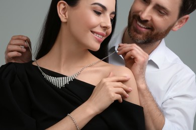 Photo of Man putting elegant necklace on beautiful woman against grey background, closeup