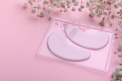 Package with under eye patches and flowers on light pink background, closeup. Cosmetic product