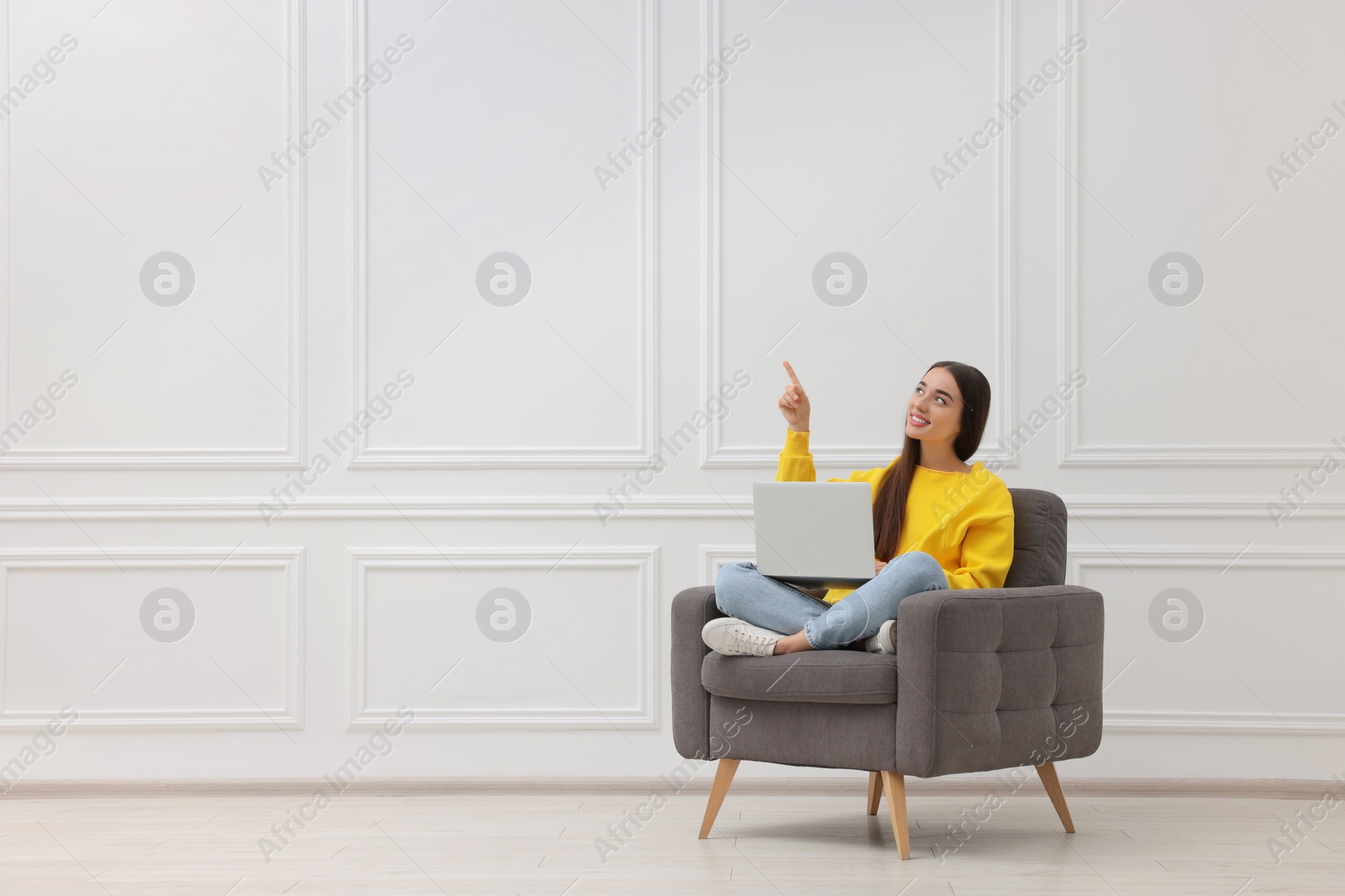 Photo of Beautiful woman with laptop pointing at something while sitting in armchair near white wall indoors, space for text